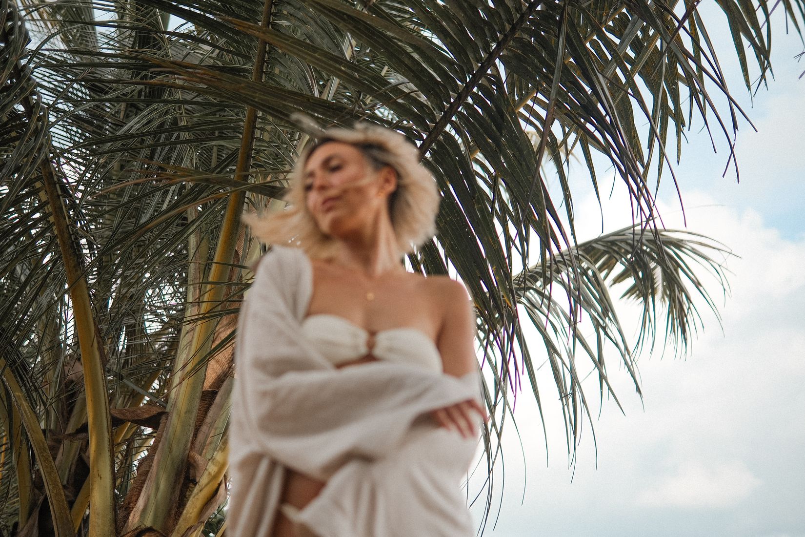 Woman in a cream bandeau and white linen shirt under palm fronds, soft focus