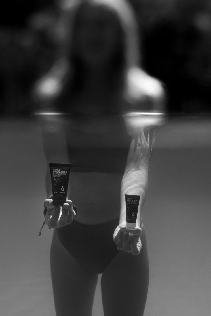 Black and white shot of a woman standing in a pool, holding a sunscreen tube in each hand