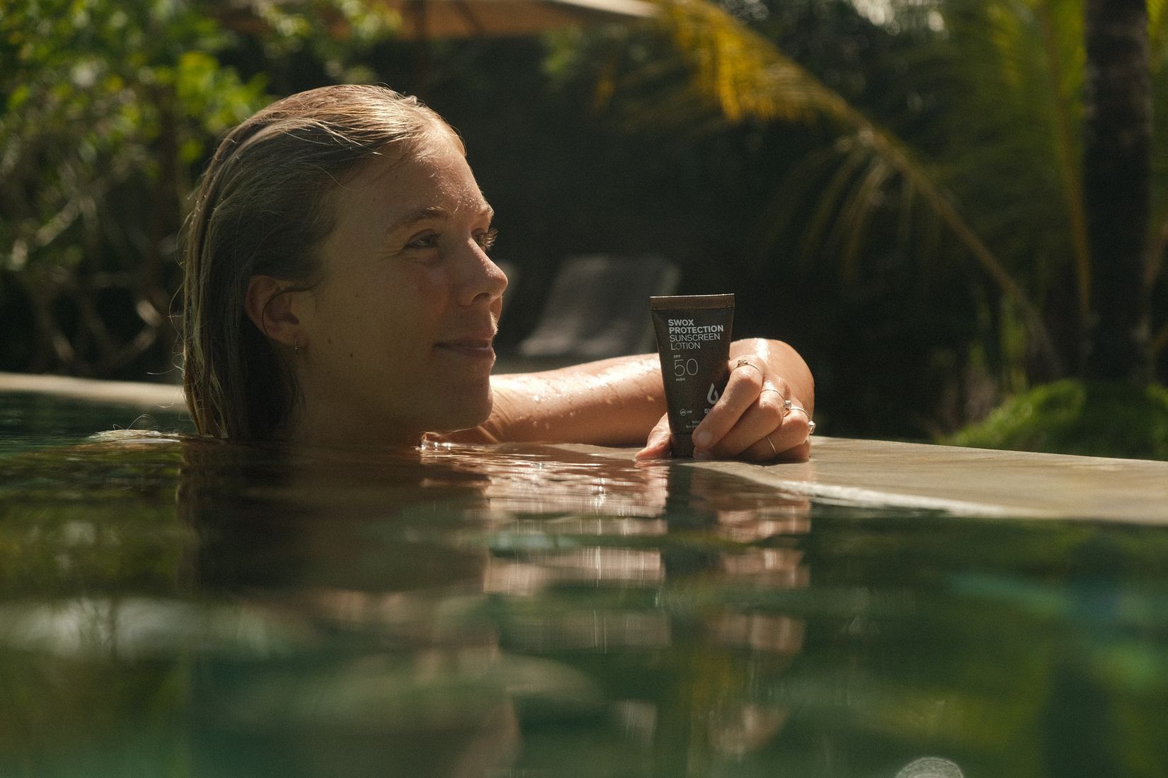 Woman in a pool resting on the edge next to a Swox sunscreen tube, smiling