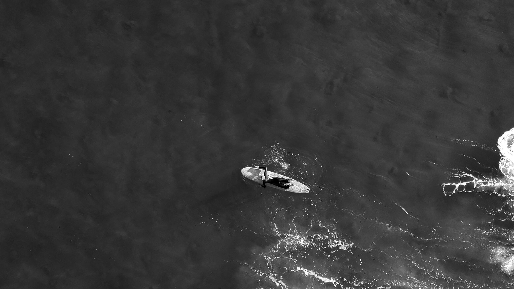 Black and white aerial view of a surfer paddling on dark water beside breaking foam
