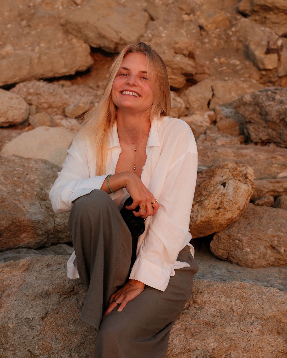 Jule smiling, holding a camera while sitting on warm-toned rocks