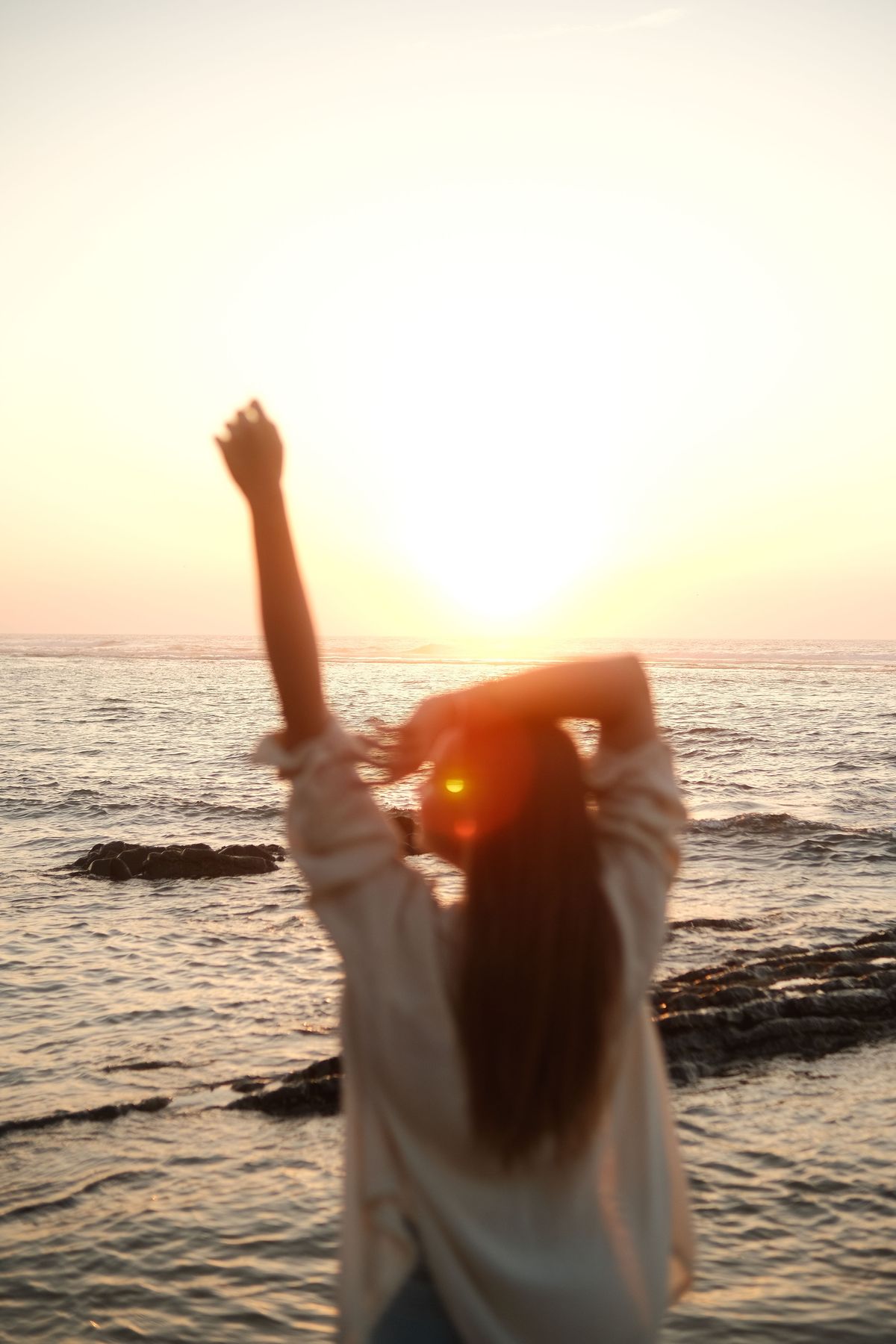 Woman seen from behind with one arm raised toward the setting sun over the sea