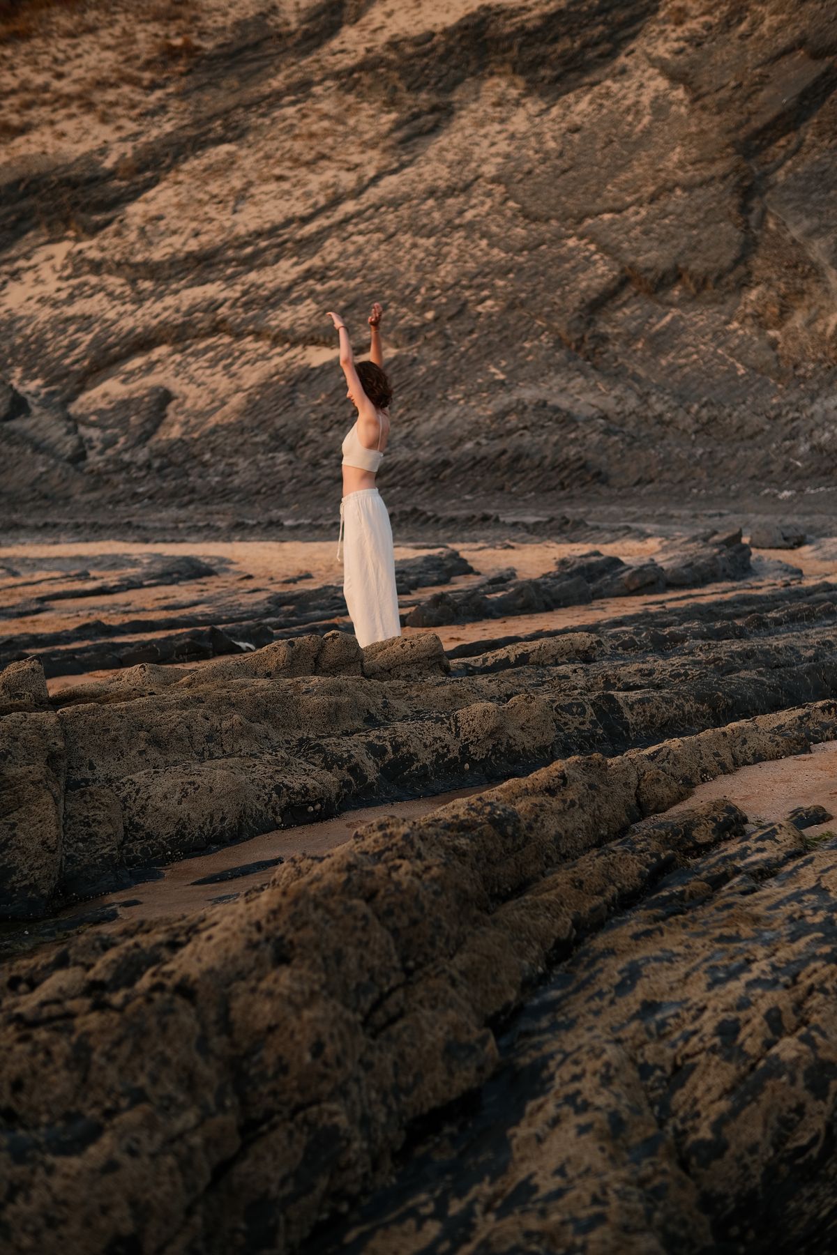 Woman in white standing with one arm raised on dark layered rocks