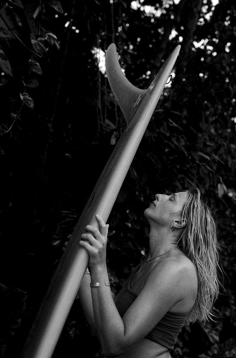 Black and white shot of a woman holding a longboard upright, looking up at the fin