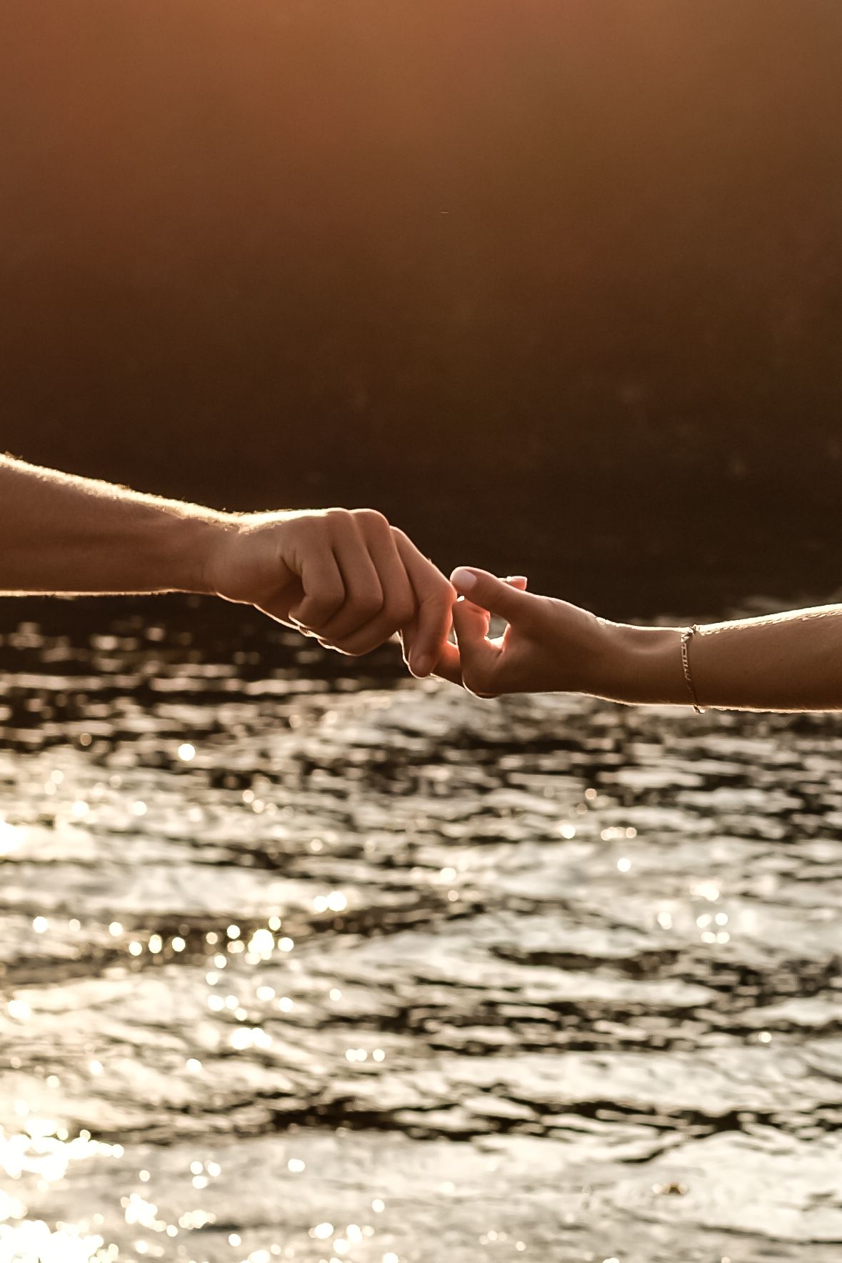 Two hands holding on to each other over glittering water at sunset