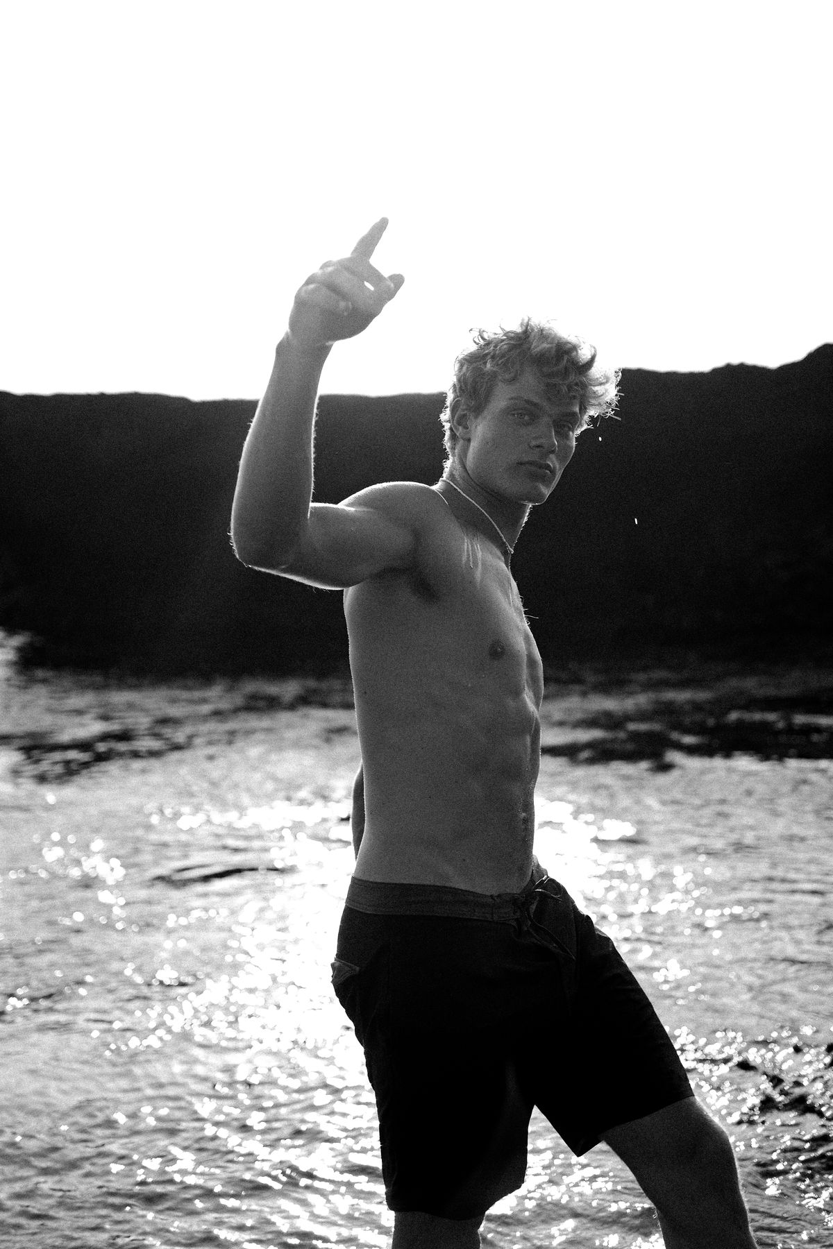 Black and white shot of a shirtless man in the water, one arm raised, pointing up