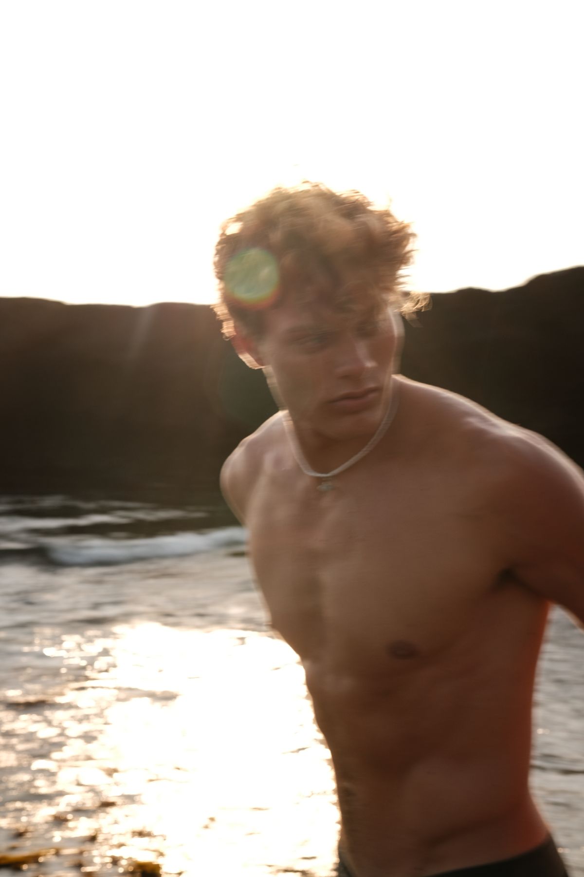 Shirtless man with curly hair walking through the water in backlight