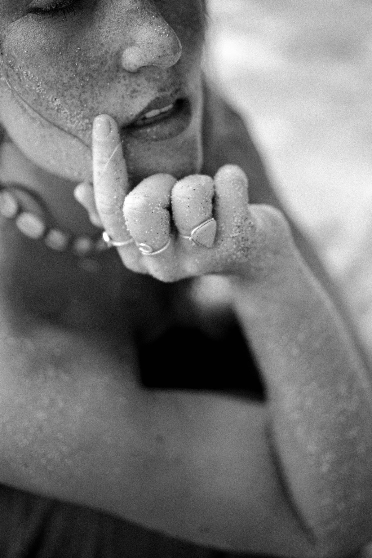 Black and white close-up of a hand with rings at a woman's lips, sand on her skin