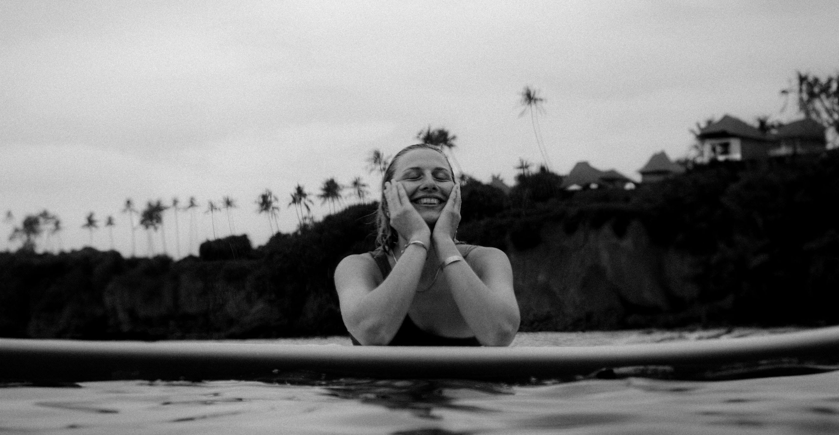 Black and white shot of a woman leaning on a surfboard in the water, hands on her cheeks, smiling with her eyes closed, palm trees and villas behind