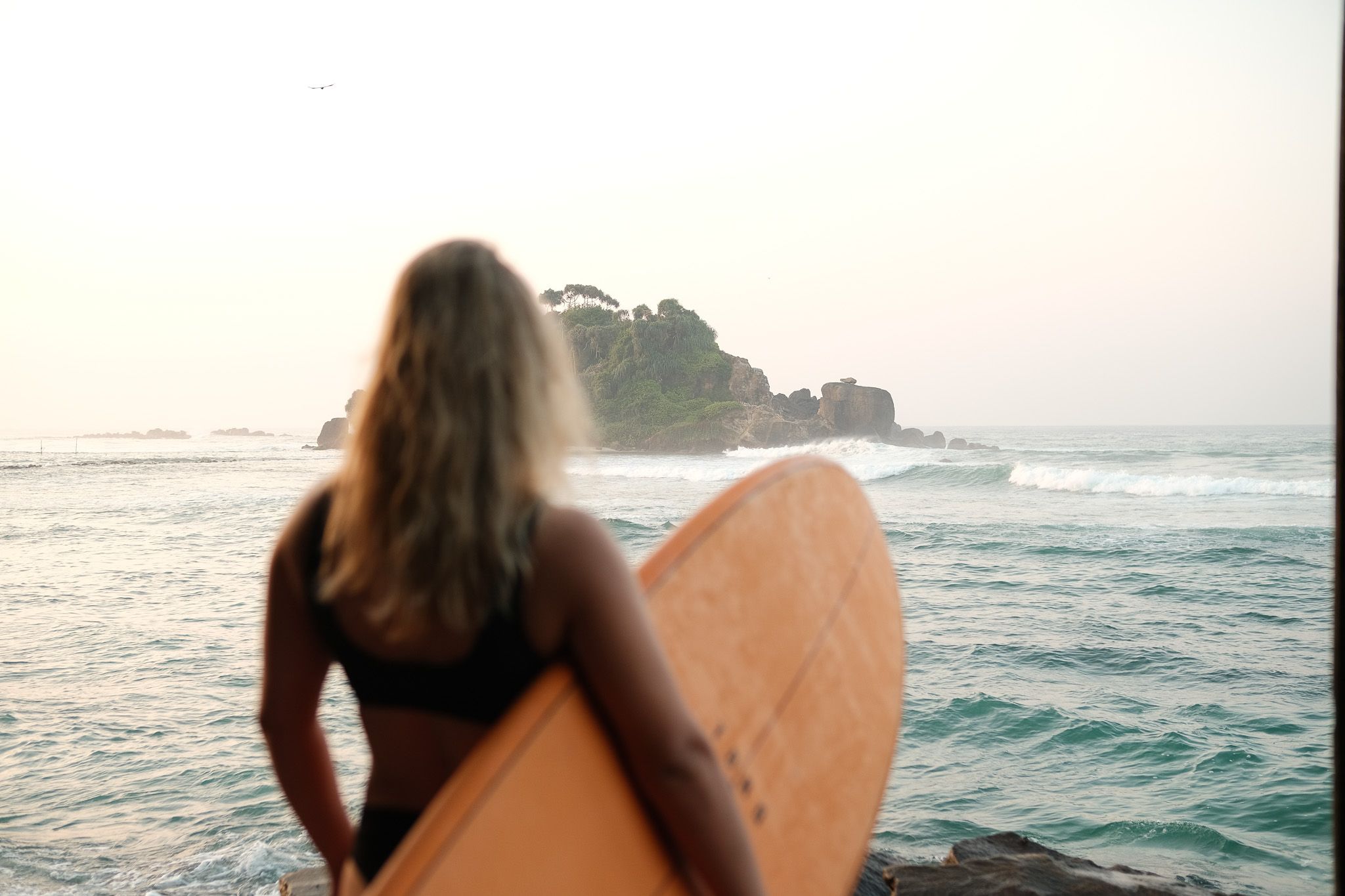 Woman holding a surfboard looking out at the sea and a small island