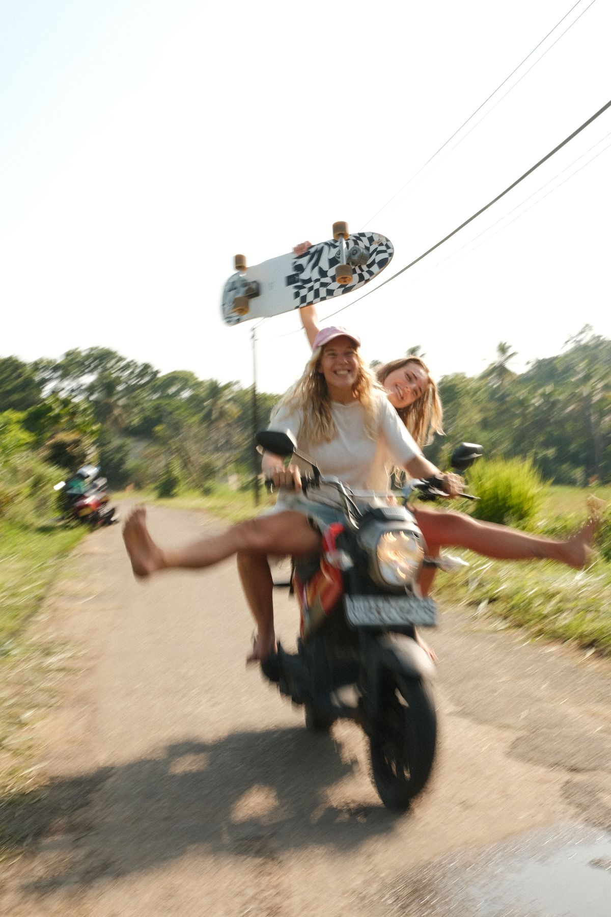 Two women on a scooter, one holding up a skateboard, legs stretched out