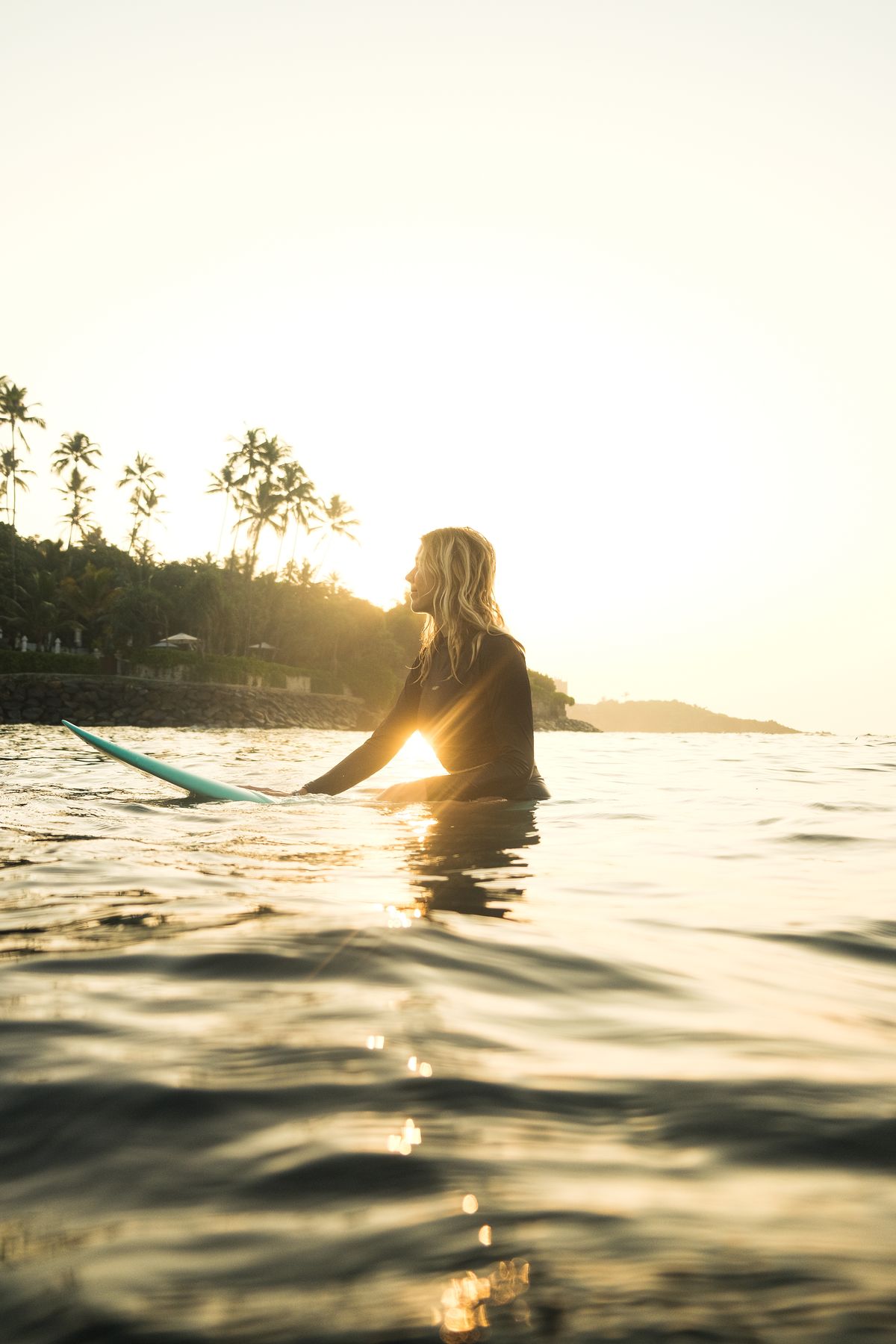 Woman holding her surfboard in the water, backlit by the low sun