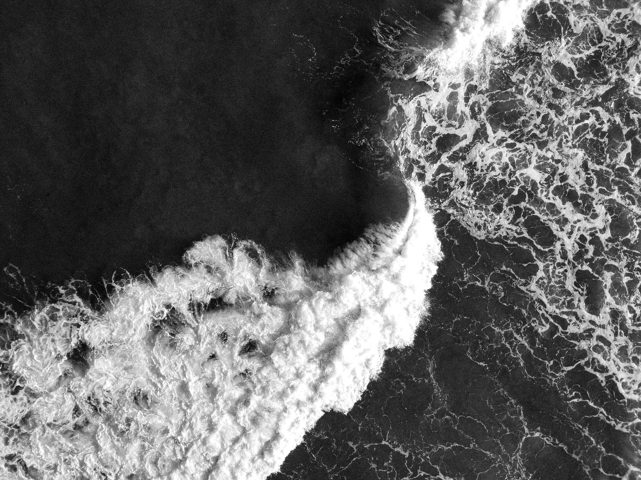 Black and white drone shot of a wave breaking into white foam