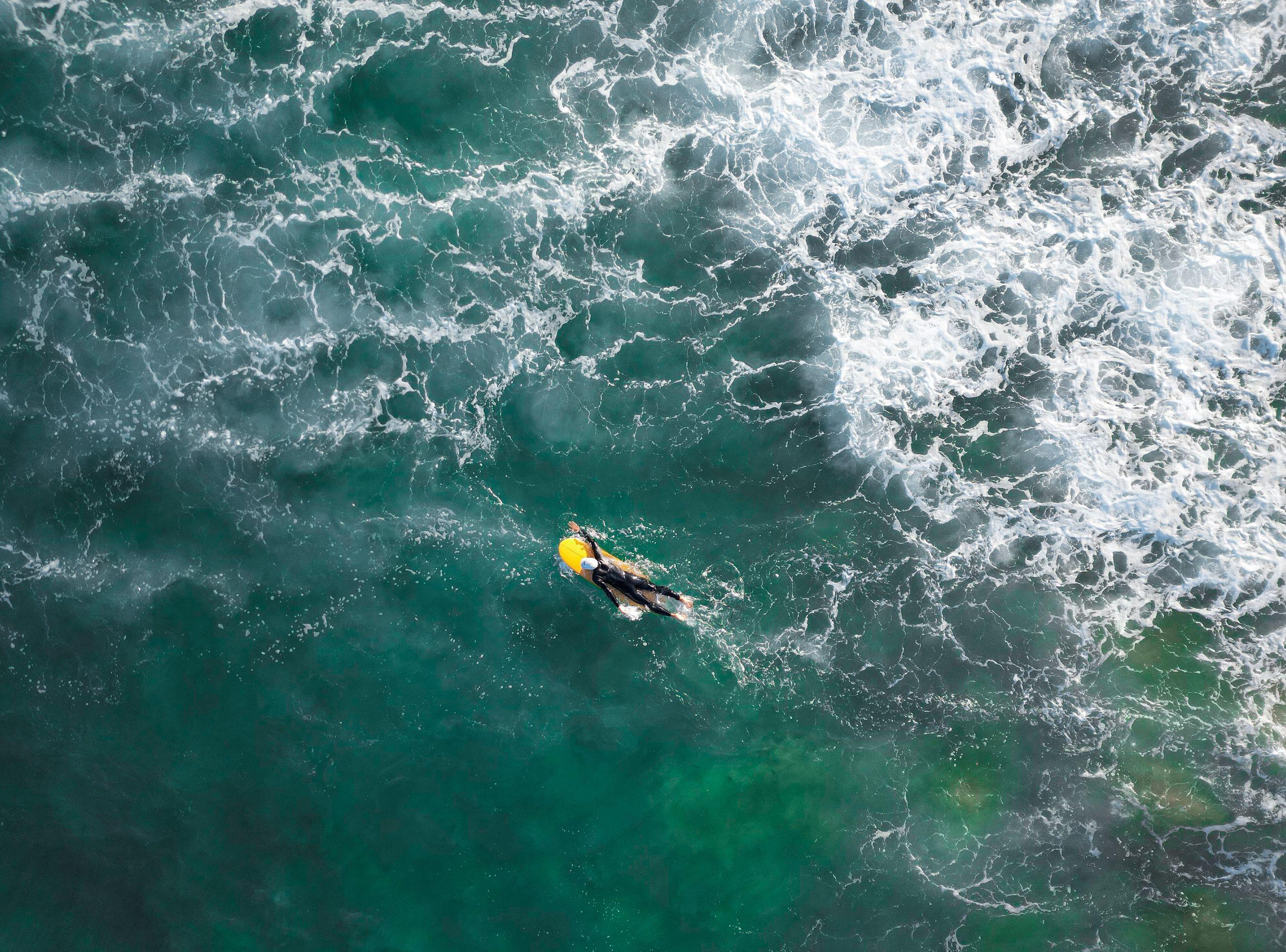 Drone shot of a surfer paddling through white water on a teal ocean