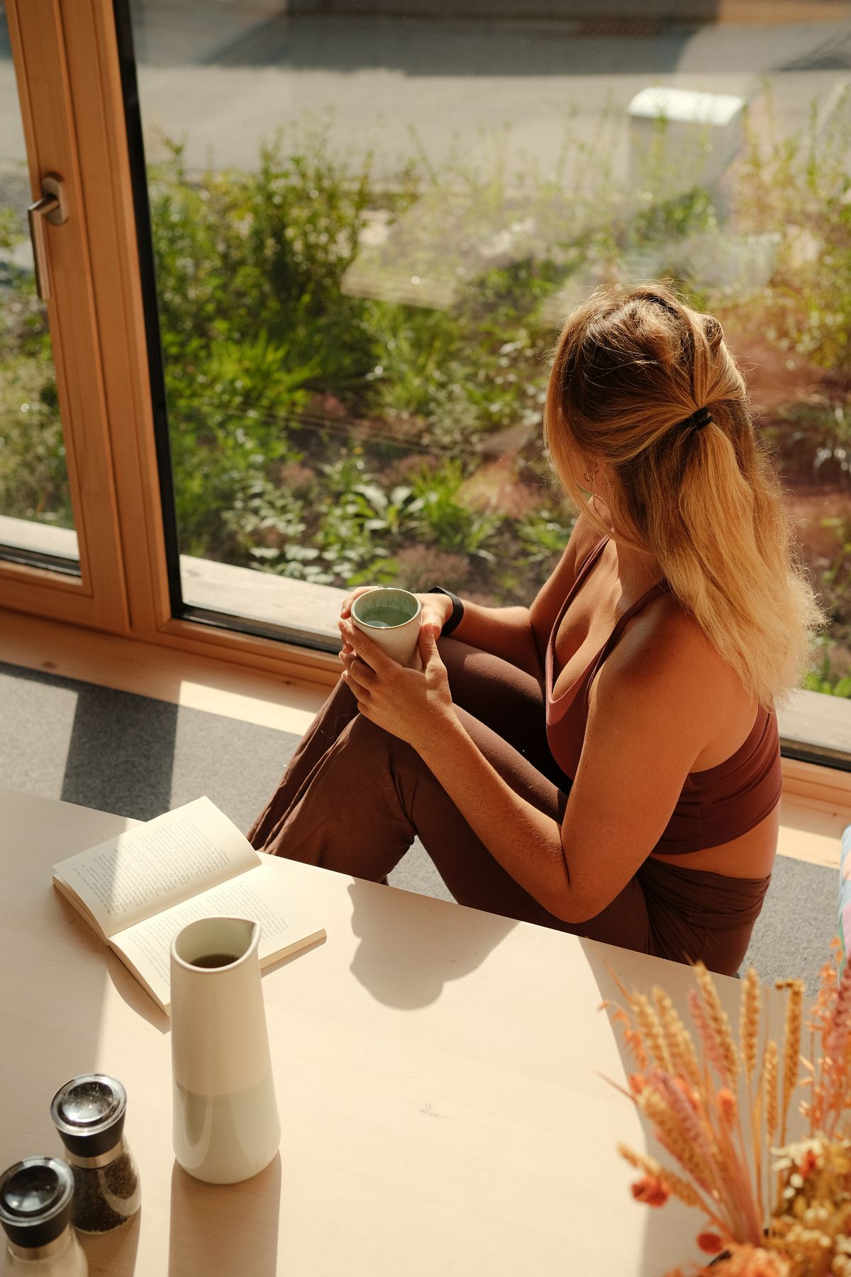 Woman with a cup sitting at the big cabin window, looking out at the garden